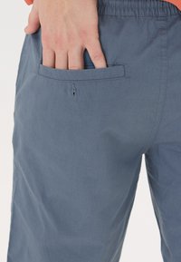 Blå shorts med en slät yta, featuring ett elastic waistband och en bakficka. En hand vilar i fickan, vilket visar på avslappnad funktionalitet.