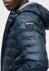 Κοντινό πλάνο ενός ατόμου που φοράει μπουφάν φουσκωτό σε ναυτικό μπλε χρώμα με κουκούλα και κεντημένη ετικέτα Blauer στο πάνω μέρος του μανικιού.