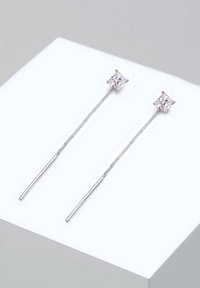 Paire de boucles d'oreilles en argent à finition filaire avec des pierres précieuses taillées en carré transparentes au sommet, présentées sur une surface blanche.