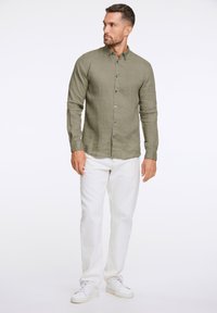 Camisa de lino verde olivo con botones y mangas largas, combinada con vaqueros de denim blancos y zapatillas blancas, mostrando un ajuste casual.