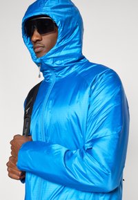 Modra izolirana jakna s kapuco, ki ima gladko, sijočo teksturo in zadrgo. Oblečena z temnimi sončnimi očali in črnim nahrbtnikom.