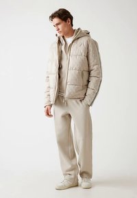 Beige-tikkitakki, jossa tekstuuri ja huppu sekä valkoiset raidat. Yhdistetty samansävyisiin beigeihin housuihin ja valkoisiin kenkiin.