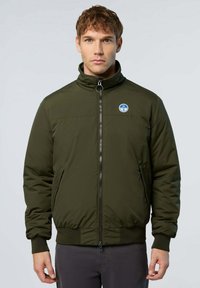 Uomo che indossa una giacca bomber verde oliva con zip e un piccolo logo tondo blu e bianco sul lato sinistro del petto, in piedi su uno sfondo semplice.