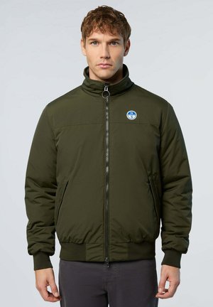 Man gekleed in een groene bomberjack met rits, met een klein rond blauw en wit logo op de linkerborst, staande tegen een effen achtergrond.