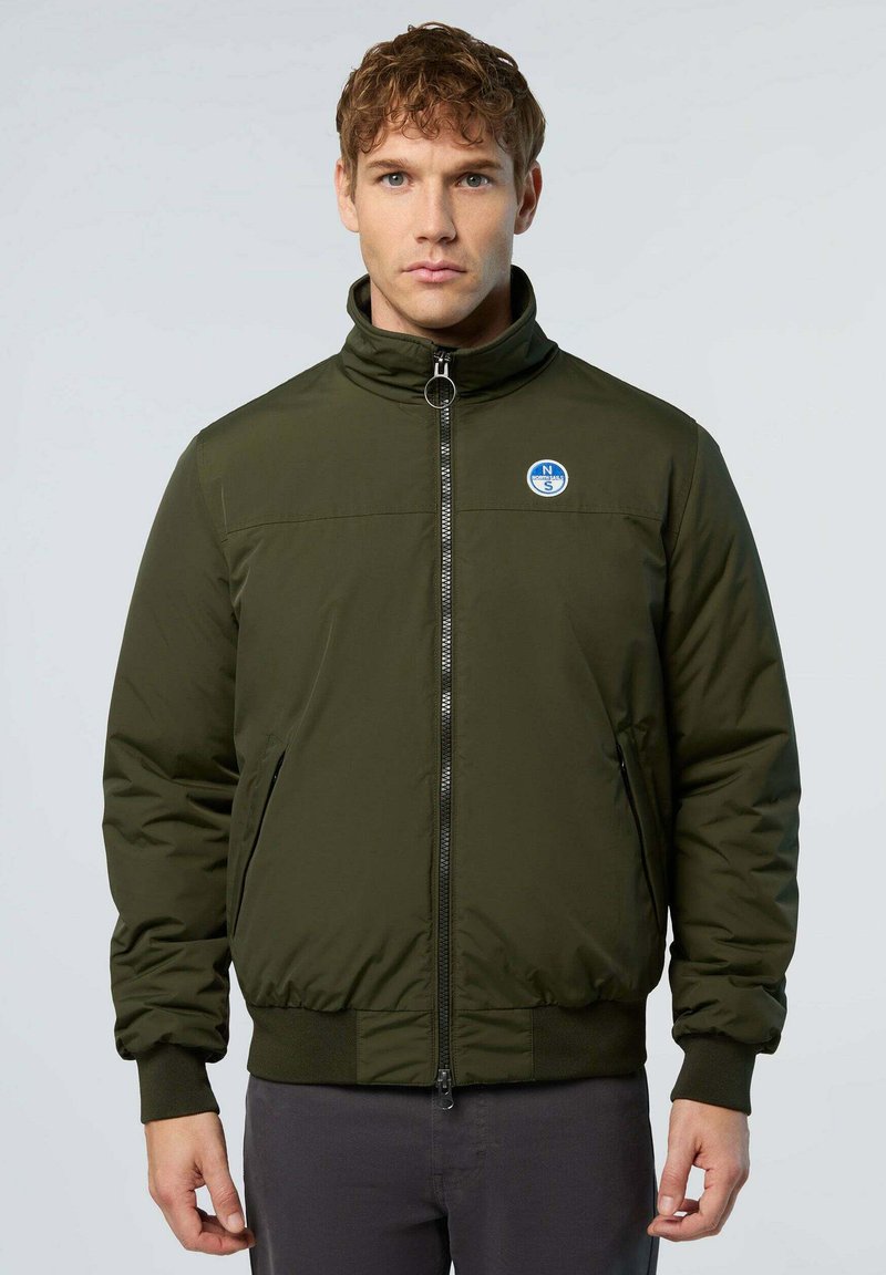 Uomo che indossa una giacca bomber verde oliva con zip e un piccolo logo tondo blu e bianco sul lato sinistro del petto, in piedi su uno sfondo semplice.