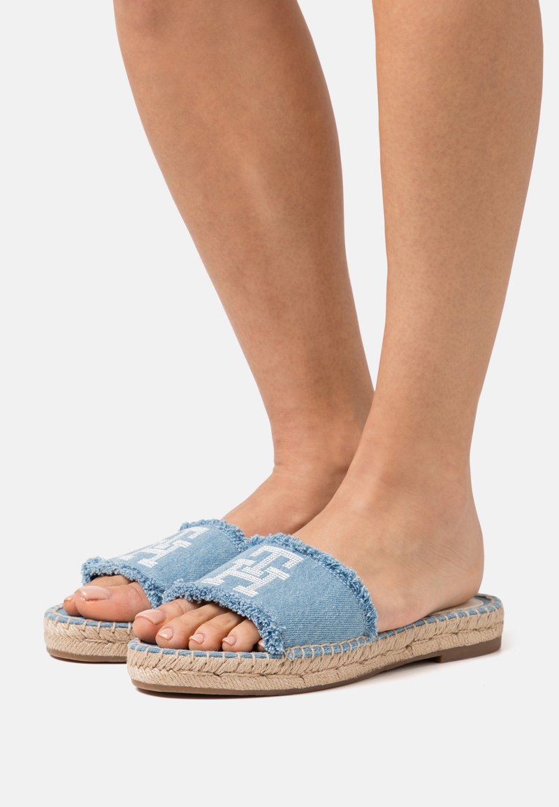 Jambes féminines portant des sandales à glissière en denim bleu avec broderie blanche et semelles en corde tressée sur un fond clair uni.