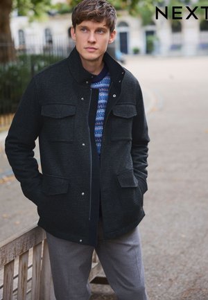 Jeune homme portant une veste noire texturée par-dessus un pull à motifs, debout dehors sur un chemin avec les mains dans les poches.