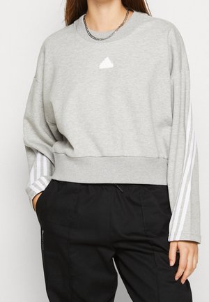 Nainen, jolla on harmaa pitkähihaisen collegepaita, jossa valkoiset raidat hihoissa ja pieni logo, yhdistettynä mustiin housuihin ja ketjukaulakoruun.