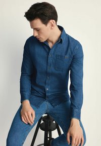 Niebieska koszula z denimu z kołnierzykiem zapinanym na guziki, długimi rękawami i detalami kieszeni. Połączona z dopasowanymi niebieskimi dżinsami.