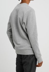 Persona vistiendo un sudadera de manga larga gris claro sin estampado y pantalones oscuros de rayas finas, de pie de espaldas sobre un fondo liso.