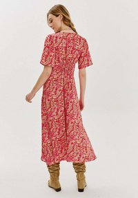 Květinové maxi šaty s krátkým rukávem, s vzorem v odstínech růžové, červené a žluté, s vypasovaným pasem a splývavou siluetou. Nosené s béžovými kozačkami.