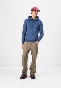 Sudadera azul con un bolsillo frontal, combinada con pantalones beiges y zapatillas negras y rojas. El modelo lleva una gorra rosa con una banda negra.