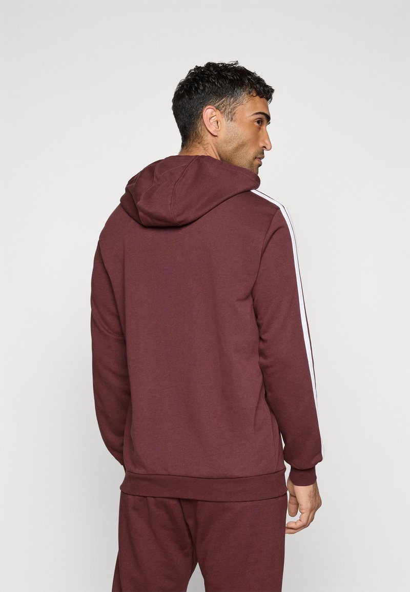 Homme de dos portant un sweat à capuche bordeaux et un pantalon assorti avec des rayures blanches le long des manches, sur fond uni.