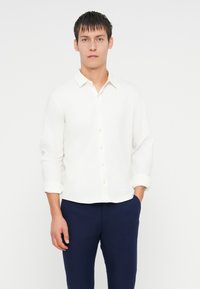 Camicia bianca con colletto, maniche lunghe arrotolate ai polsini, indossata con pantaloni navy scuri. Texture liscia, design semplice.