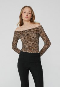 Žena v velkých kruhových náušnicích, s leopardím potiskem na topu s odkryté ramena a dlouhými rukávy, a v černých vysokých kalhotách stojí před jednobarevným pozadím.