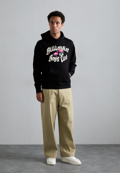 Sweat à capuche noir avec le texte "Billionaire Boys Club" et des graphismes de flamant rose, associé à un pantalon large beige et des baskets blanches.