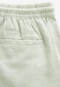 Tissu texturé vert clair avec une taille élastique et un petit détail de poche arrière, présentant un motif tissé subtil.