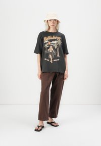 Svart grafisk t-skjorte med et retro Billabong-design, kombinert med løse brune bukser og svarte flip-flops; modellen har på seg en lys bøttehatt.