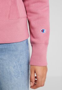 Sweatshirt crewneck rose avec des poignets côtelés, présentant un logo brodé sur la manche. Associé à un jean bleu délavé montrant des détails usés.