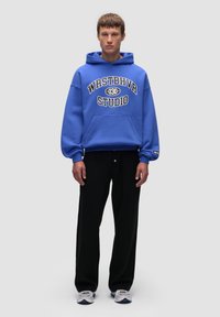 Niebieska bluza z kapturem z białym i czarnym napisem z przodu, zestawiona z luźnymi czarnymi spodniami i białymi trampkami z niebieskimi akcentami.