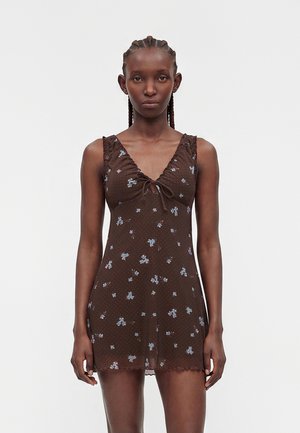 Frau mit geflochtenem Haar trägt ein ärmelloses braunes Mini-Kleid mit blauen Blumenmustern und einem vorderen Bindeband.
