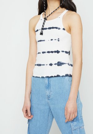 Person trägt ärmelloses weißes Tanktop mit horizontalen schwarzen Batik-Streifen und hellblaue Jeans-Cargohose, steht vor weißem Hintergrund.