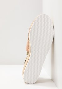 Semelle d'une sandale espadrille beige avec une semelle en caoutchouc blanc rainuré, reposant verticalement contre un mur et une surface blancs.