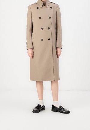 Beżowy dwurzędowy trencz z czarnymi guzikami, noszony na białych skarpetkach i czarnych błyszczących mokasynach, widoczny od ramion do połowy łydki.