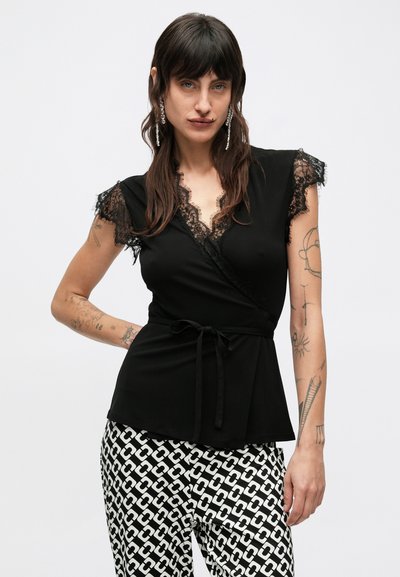 Czarna bluzka kopertowa z krótkimi koronkowymi rękawami, głębokim dekoltem w kształcie litery V wykończonym koronką oraz wiązaniem w talii. Połączona z czarno-białymi spodniami w wzory.