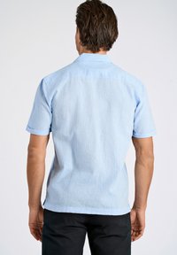 Vaaleansininen lyhythihainen paita, jossa on teksturoitu pinta, nappikaulus ja käännetyt hihat, yhdistettynä tummiin housuihin, näkyy takaapäin.