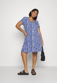 Vestido floral azul com mangas curtas, decote em V e barra evasê. O tecido tem uma textura ligeira. Combinado com sandálias pretas e uma mala preta.