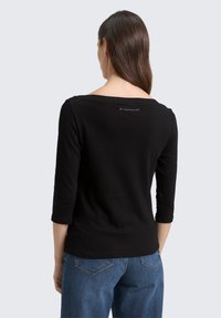 Schwarz langärmliges Shirt mit weitem Ausschnitt, weichem Material und einem Logo-Detail auf dem Rücken; kombiniert mit blauen Jeans.
