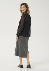 Schwarze, transparente Bluse mit Polka-Dots und langen Ärmeln, kombiniert mit einem grauen, knielangen Rock mit seitlichen Schlitzen und Taschen. Schwarze, hochhackige Mules.