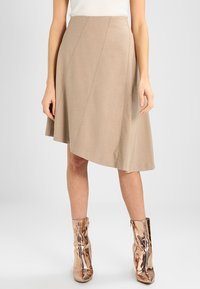 Jupe asymétrique longueur genoux en tissu beige avec des coutures diagonales ; présente une texture lisse et une légère drape. Associée à des bottes métallique doré rose.