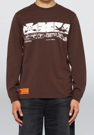 Persona che indossa una maglietta a maniche lunghe marrone con una grafica astratta bianca e il testo "Forgotten Times Spiral Sight" sul petto.