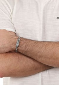 Pulsera de cadena de plata con un cierre rectangular texturizado, llevada en una muñeca sobre un fondo de tela texturizada de color claro.