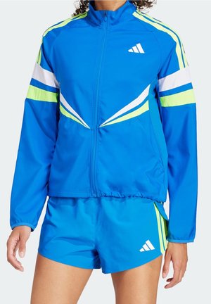 Person iført blå atletikjakke og matchende shorts med hvide og limegrønne striber og Adidas-logoer, stående med afslappede arme.