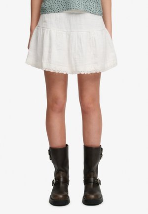 Une personne porte une mini-jupe blanche plissée avec des bordures ajourées et des bottes en cuir marron foncé mi-mollet avec des boucles, debout devant un fond blanc.