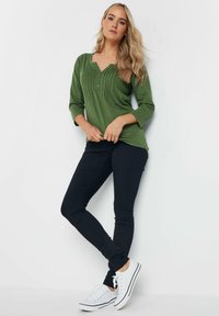 Top verde a maniche lunghe con scollatura a V e dettagli testurizzati sul davanti, abbinato a jeans skinny neri e sneaker bianche.