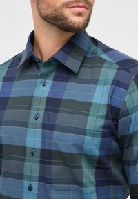 Uomo che indossa una camicia a quadri blu e verde con taschino frontale, mostrato dalla zona del collo fino a metà torace su sfondo bianco.