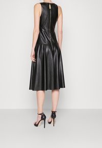 Kvinna som bär en ärmlös svart läder midi-klänning med dragkedja i ryggen och svarta ankelsandaler med hög klack mot en neutral bakgrund.