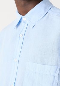 Camicia di lino azzurro chiaro con colletto a punta, bottoni bianchi e una singola tasca applicata sul lato sinistro. Tessuto testurizzato con una vestibilità casual.