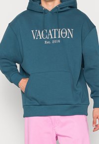 Felpa teal con tasca frontale, cappuccio con cordino e testo stampato "VACATION CLUB Est. 2016" in colore chiaro sul petto. Texture morbida.