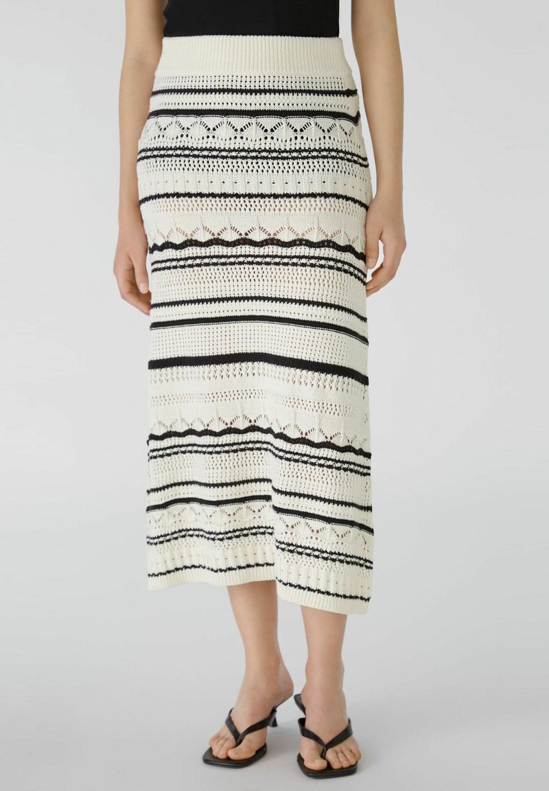 Jupe tricotée crème longueur genou avec rayures noires horizontales et motifs en dentelle, portée avec un top noir sans manches et des sandales noires à talons.