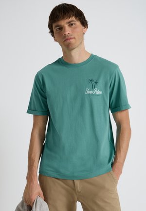 Homme portant un t-shirt bleu sarcelle "Twin Palms" et un pantalon beige, tenant un tissu gris dans la main gauche, debout devant un fond clair uni.