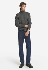 Grau gestrickter Rollkragenpullover mit gerippten Bündchen und Saum, kombiniert mit dunkelblauen Jeans und schwarzen Schuhen mit braunen Schnürsenkeln, getragen von einem männlichen Modell.