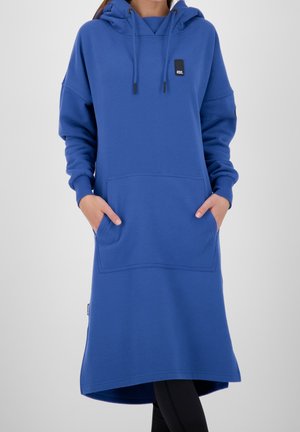 Kvinde iført en lang blå hættetrøjekjole med forlomme og sorte leggings, hænderne i lommerne, foran en ensfarvet baggrund.