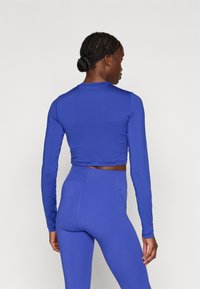 Ilgų rankovių, trumpas viršus ir aukštos talijos leggingai ryškiai mėlynos spalvos. Minkšta, tampri medžiaga su lygiu paviršiumi ir minimaliomis siūlėmis.