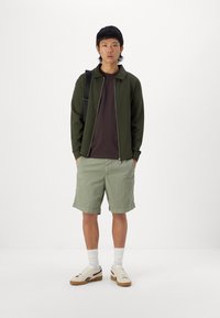 Alyvuogių žalios spalvos zip-up striukė, ruda marškinėliai, šviesiai žali šortai, balti kojinės ir grietinėlės spalvos sportbačiai su juodais akcentais. Paprastas, kasdieniškas drabužių derinys.