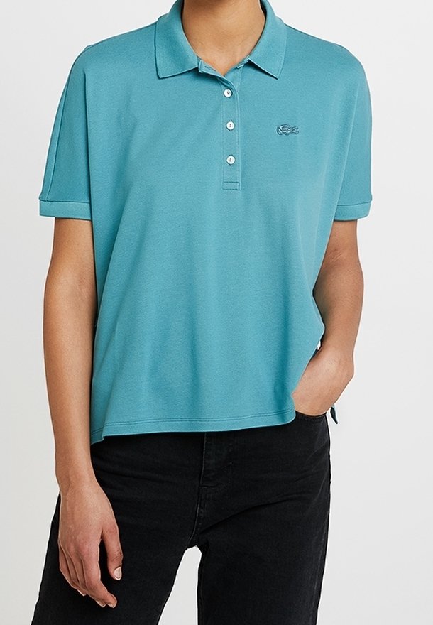 Personne portant un polo à manches courtes bleu sarcelle avec des boutons et un logo brodé, associé à un pantalon noir, main dans la poche.